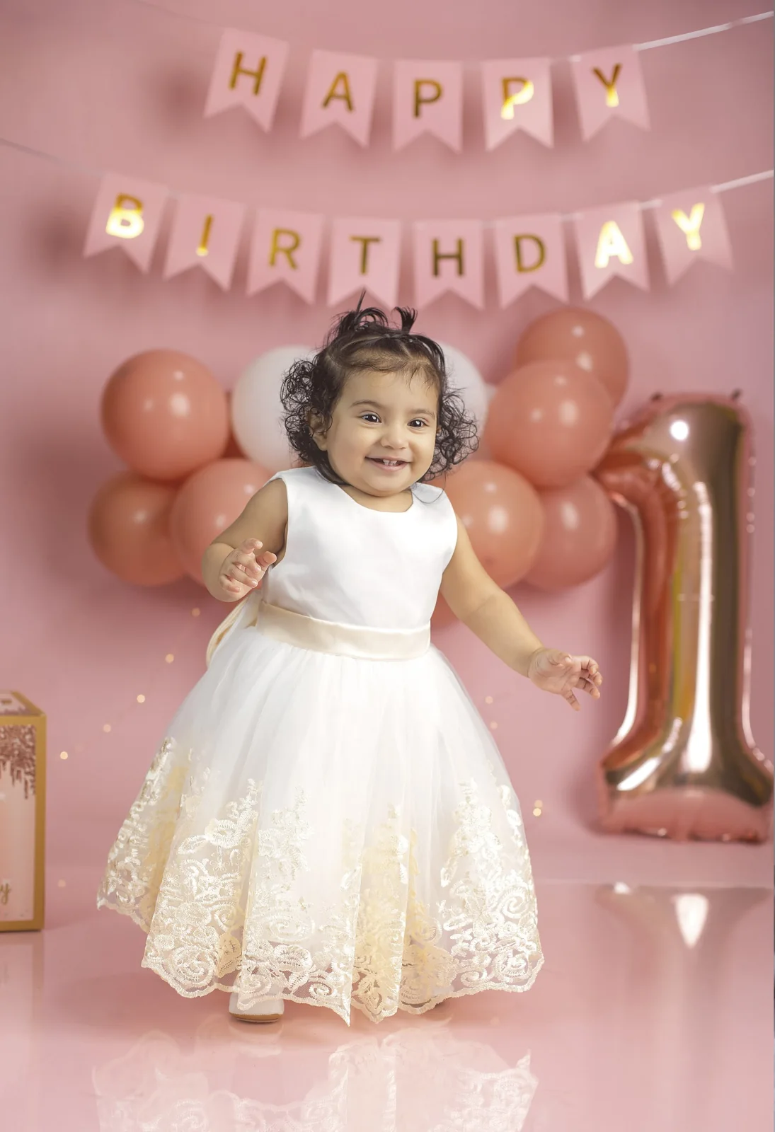 Bebé celebrando su primer cumpleaños con vestido blanco y fondo rosado